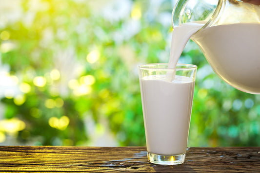 Pouring fresh milk into a clear glass on a wooden table outdoors.
