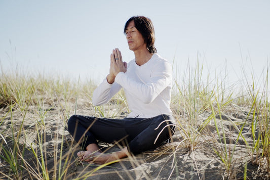 Man meditating on a sandy beach surrounded by grass.