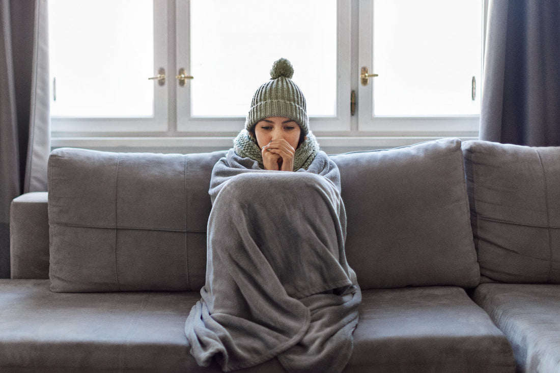 Woman wrapped in blanket sitting on couch, feeling cold.