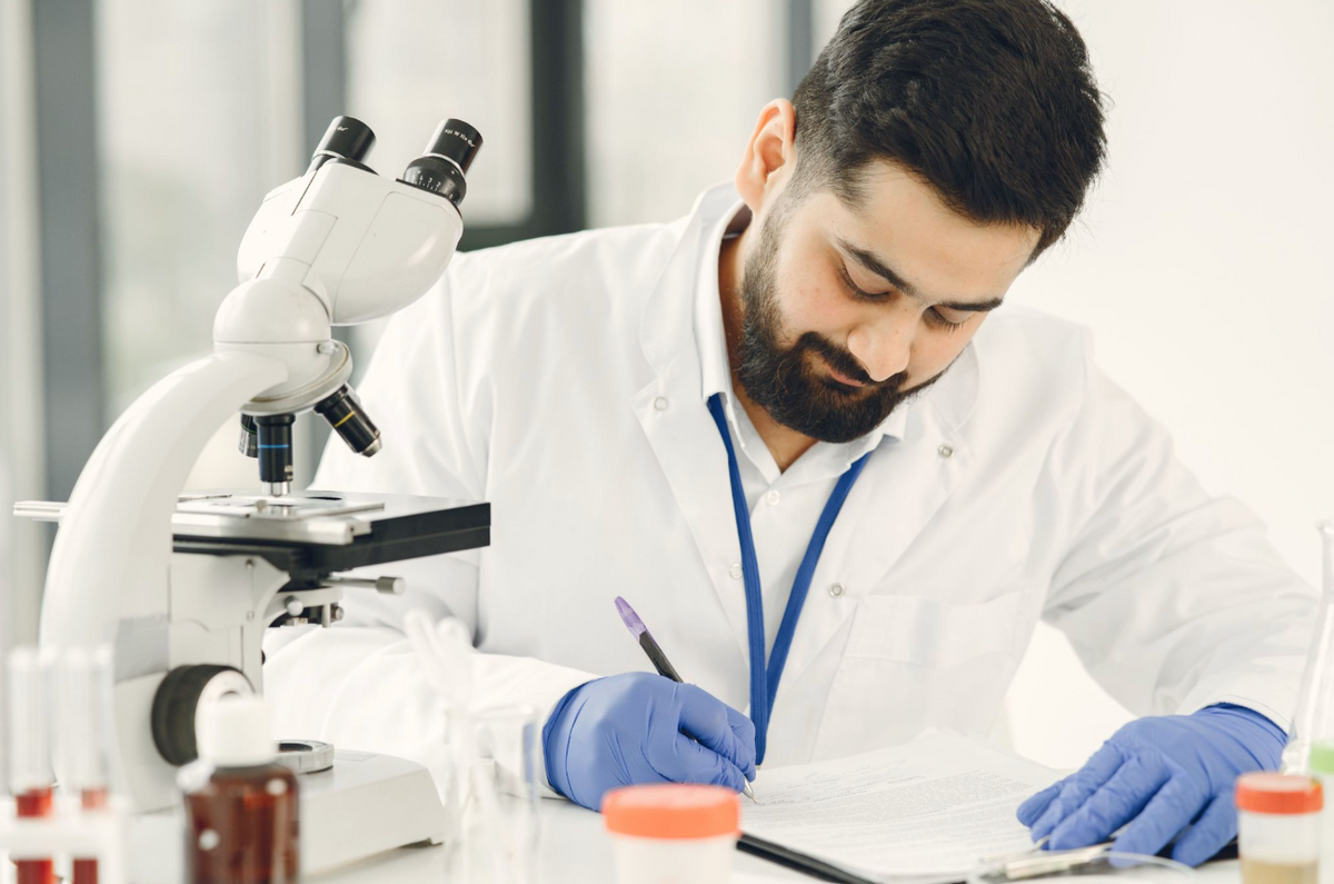 A medical professional wearing a white lab coat and blue gloves sits at a desk, looking down and writing on a clipboard with a pen, next to a microscope.