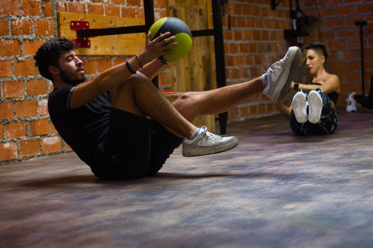 A person lies on a gym floor holding a weighted medicine ball while doing an abdominal exercise, with another person exercising in the background near a brick wall.