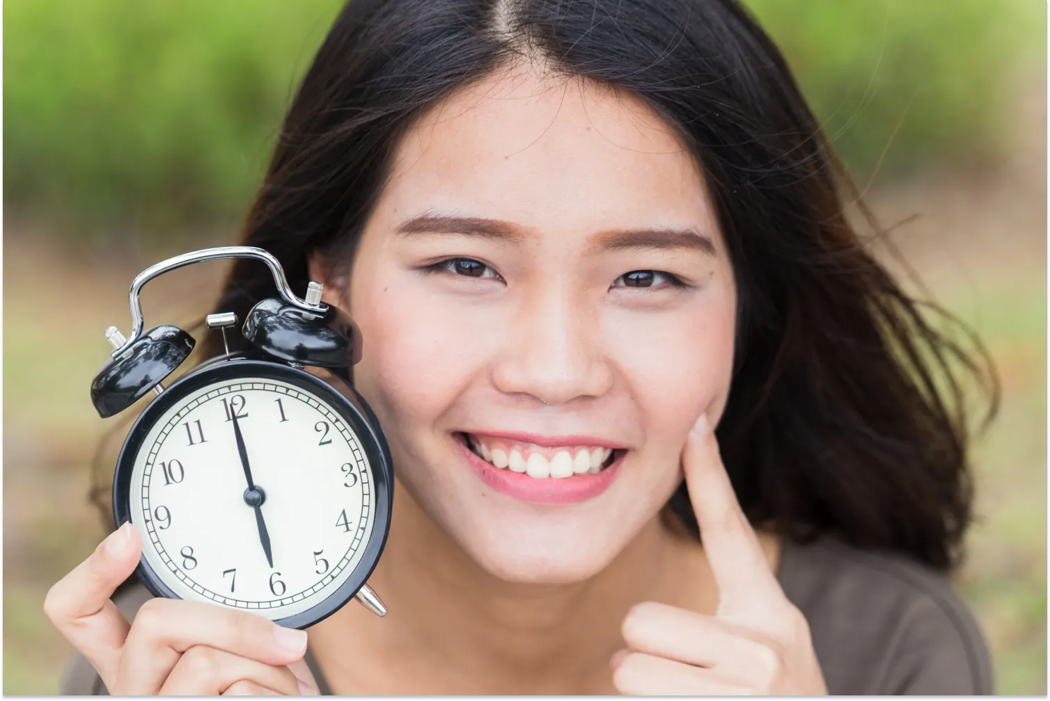 Smiling person holding an alarm clock outdoors.