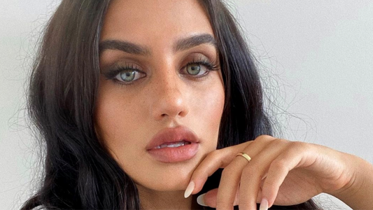 Close-up of a person with long dark hair and green eyes, posing thoughtfully with hand near face.