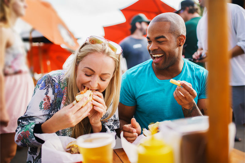 Happy couple enjoying food at an outdoor market under red umbrellas.