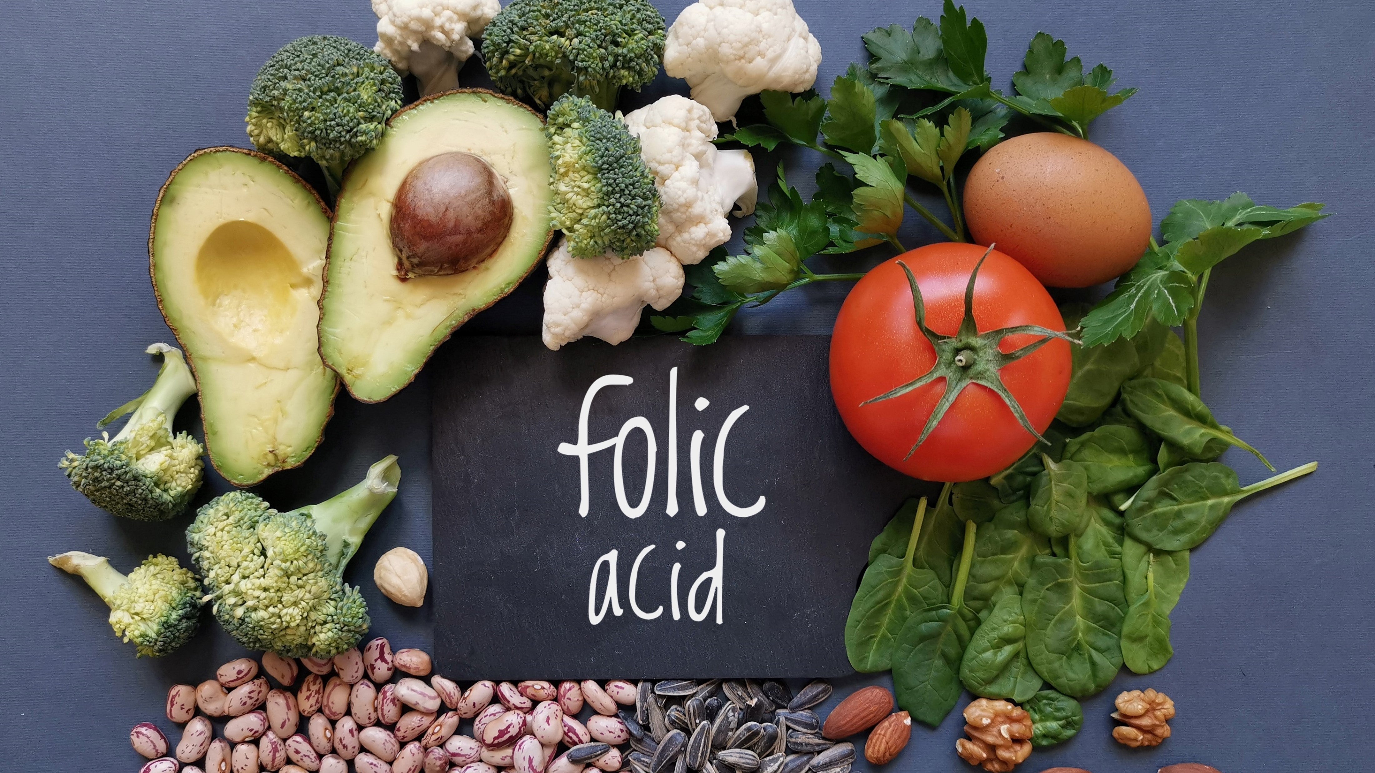 flat lay of foods rich in folic acid including avocado, broccoli, tomato, beans, nuts and egg with chalkboard reading 'folic acid'