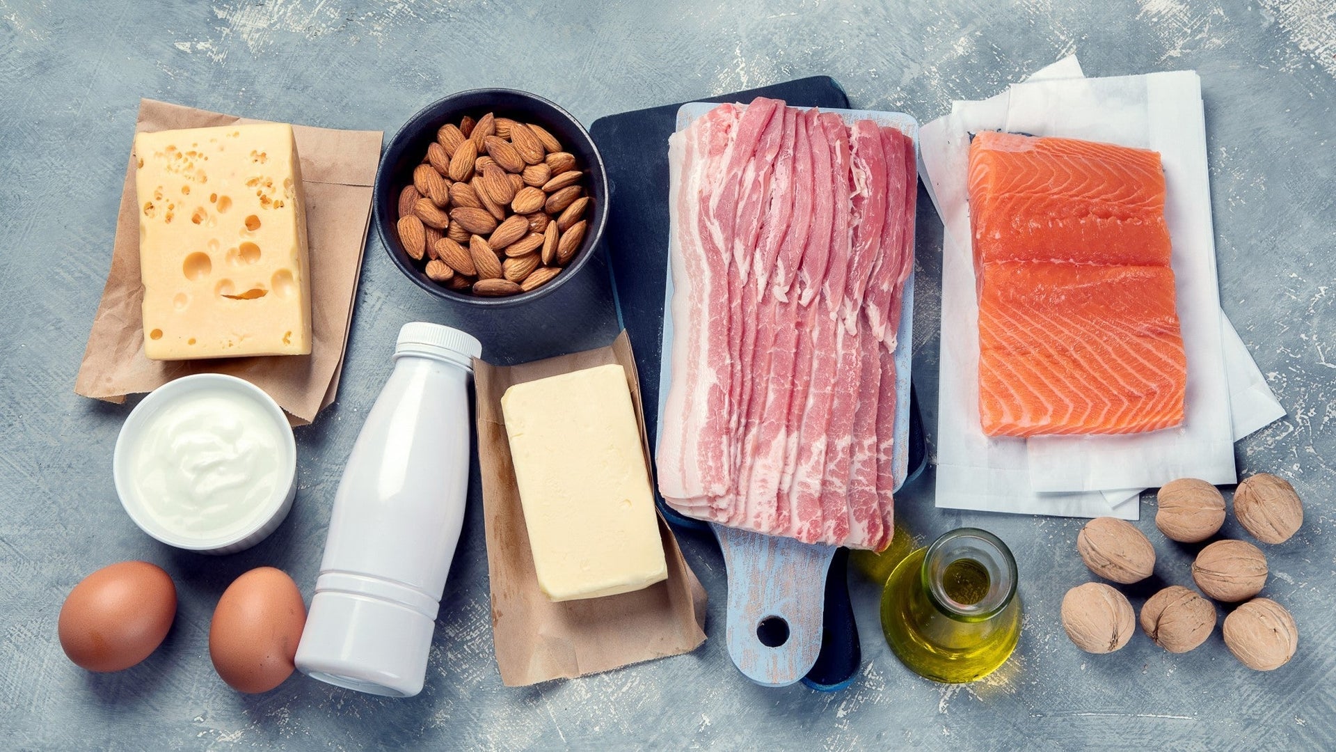 Keto-friendly foods: cheese, almonds, bacon, salmon, yogurt, eggs, oil, and nuts on a table.